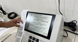 #PraTodosVerem: Na fotografia aparece a mão de uma pessoa, segurando um documento, a frente de u...