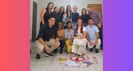 #PraTodosVerem: Na fotografia homens e mulheres sorrido para a câmera, em uma sala. No centro do...