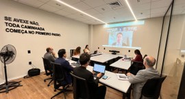 Na fotografia aparece várias pessoas sentadas, em torno de uma mesa retangular. Na projeção, apa...