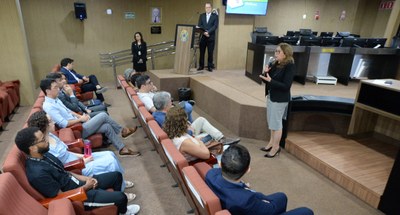 #PraTodosVerem: Na fotografia aparece uma mulher em pé, falando em um microfone, à frente de pes...
