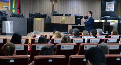 #PraTodosVerem: Na fotografia aparece uma homem em pé, falando em um microfone, à frente de pess...