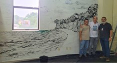 #PraTodosVerem: Na fotografia aparecem três homens em pé, ao lado de um mural artístico que repr...