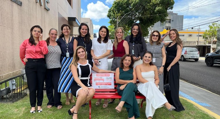 Banco Vermelho é instalado no TRE-PB para reflexão contra feminicídio #PraTodosVerem: Na fotografia aparece um grupo de mulheres em torno do Banco vermelho instalado ...