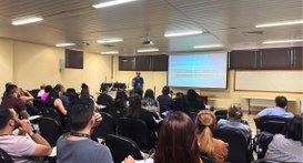 #PraTodosVerem: Na fotografia aparece um grupo de pessoas sentadas assistindo uma palestra em fr...
