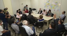 Pessoas numa sala de reunião, em volta de uma mesa, e uma outra de pé filmando.