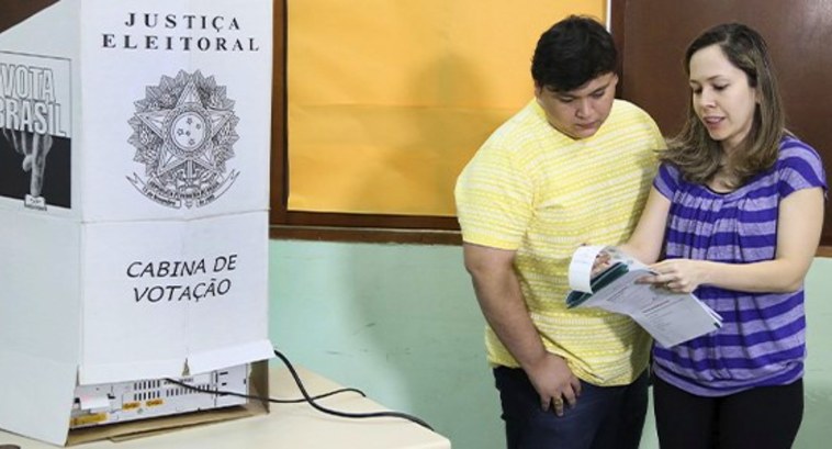 Dia do Voluntariado: voluntários superam convocados na JE #PraTodosVerem: Na fotografia, um homem e uma mulher em uma sala. À esquerda, uma cabine de vota...