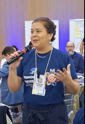 #PraTodosVerem: Na fotografia aparece, em primeiro plano, uma mulher falando em um microfone.