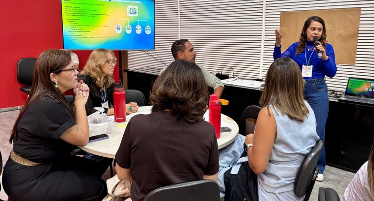 #PraTodosVerem: Na fotografia aparece uma mulher de pé, com blusa azul e microfone, falando para...