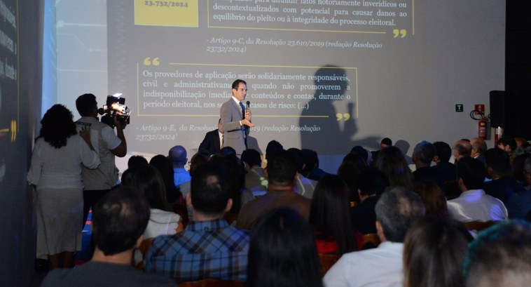 Juiz Bruno Teixeira fala sobre IA em evento sobre eleições 2024 #PraTodosVerem: Na fotografia aparece em destaque um homem segurando um microfone em cima de um ...