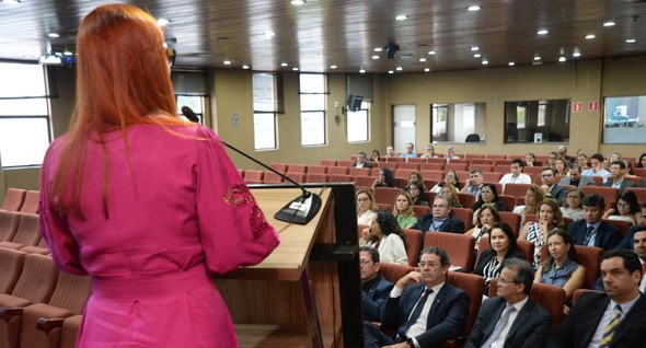 #PraTodosVerem: Mulher de vestido rosa discursa para plateia em auditório de poltronas laranjas.