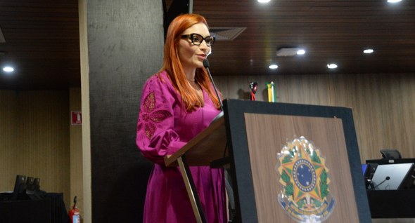 #PraTodosVerem: Mulher de cabelos ruivos e vestido rosa fala ao microfone em tribuna com brasão ...