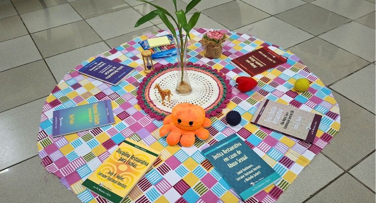 Justiça Restaurativa do TRE-PB avança agenda no interior e na sede #PraTodosVerem: Na fotografia aparece um tecido colorido no chão em formato circular, com livros...