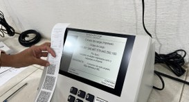 #PraTodosVerem: Na fotografia aparece a mão de uma pessoa, segurando um documento, a frente de u...