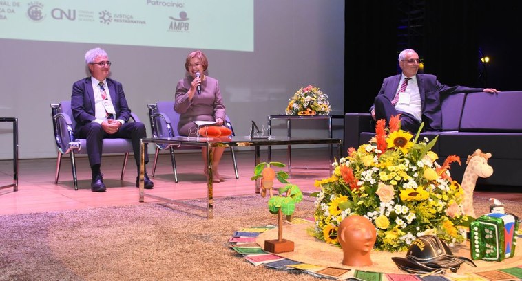 Painel JR #PraTodosVerem: Na fotografia aparecem três pessoas sentadas no palco do Congresso, tendo a medi...