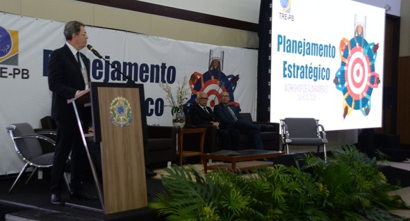 #PraTodosVerem: Na fotografia aparece um homem em cima de um palco falando em um microfone.