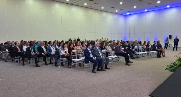 #PraTodosVerem: Na fotografia aparecem pessoas sentadas em cadeiras em uma sala com o pé direito...