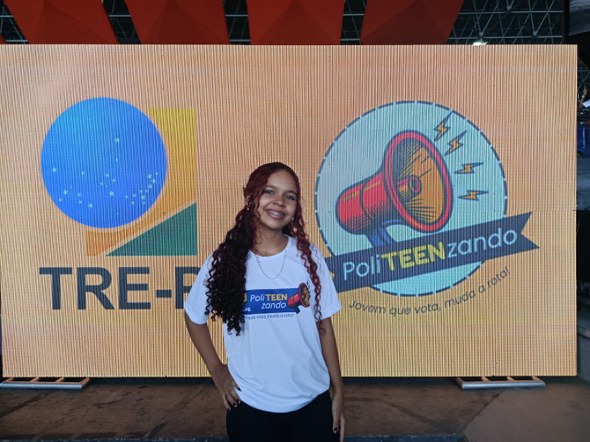 #PraTodosVerem: Na fotografia aparece uma jovem, sorrindo e usando uma camiseta branca com uma e...