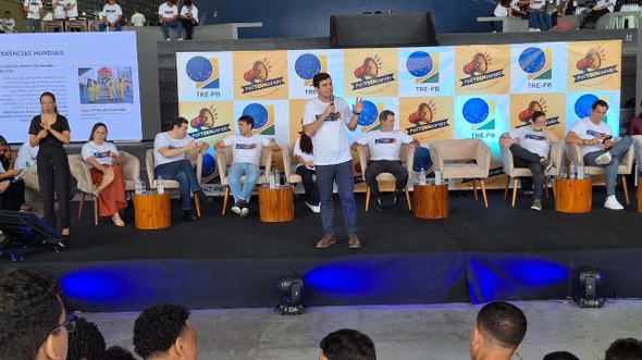 #PraTodosVerem: Na fotografia aparece um palco com um homem em pé, ao centro, vestindo uma camis...