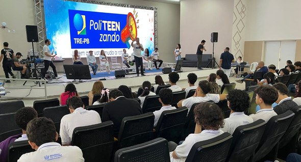 #PraTodosVerem: Na fotografia aparecem alunos sentados de costas em um auditório, assistindo a u...