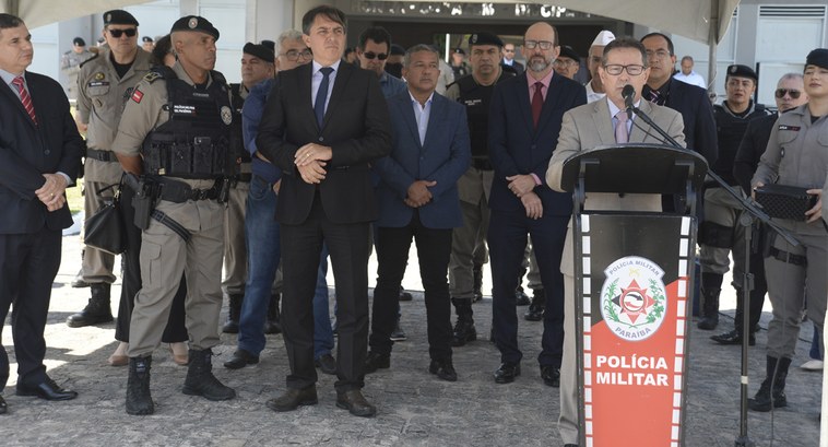 Homens de pé e um deles discursa ao microfone num púlpito a frente dos demais.