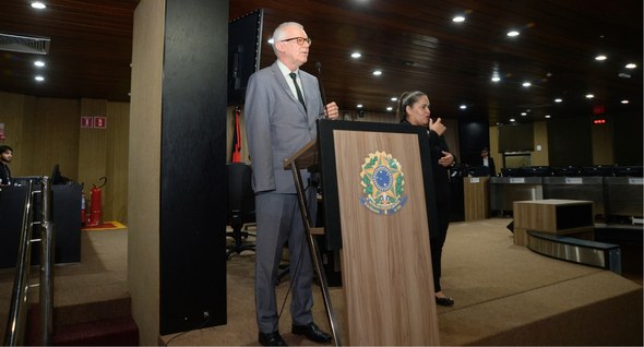 #PraTodosVerem: Na fotografia, um homem de terno fala ao microfone em um púlpito com o brasão da...