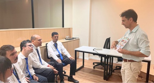 #PraTodosVerem: Na foto aparece um homem em pé que apresentando um material a quatro homens e um...