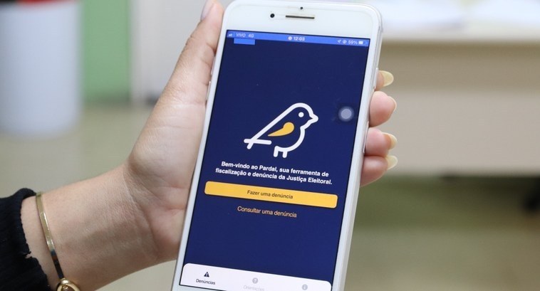 Propaganda Eleitoral irregular – denuncie pelo aplicativo Pardal #PraTodosVerem: Na fotografia aparece uma mão segurando um smartphone e na tela aparece o aplica...