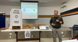 #PraTodosVerem: Na fotografia aparece um homem de pé ao lado de uma mesa contendo uma cabina de ...