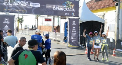 #ParaTodosVerem: Painel retangular com três pessoas ao fundo, no canto esquerdo, acenando, cada,...