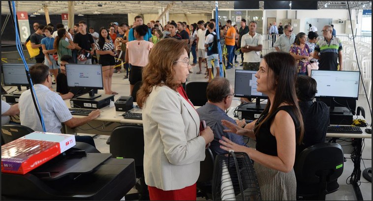 #ParaTodosVerem: Em primeiro plano, duas mulheres estão conversando de pé; no plano de fundo, pe...