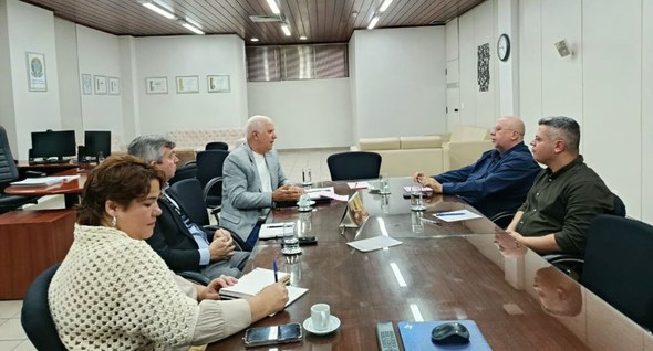 Na fotografia aparecem pessoas dialogando em uma sala de reunião.