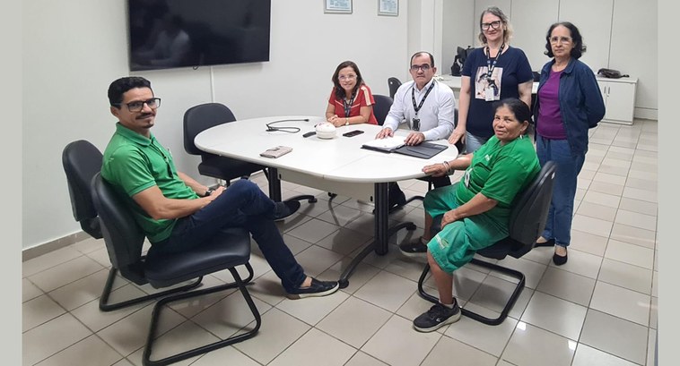 TRE-PB doa bens mobiliários à Cooperativa de Reciclagem #ParaTodosVerem: Painel retangular com pessoas reunidas em volta de uma mesa, quatro sentadas e ...