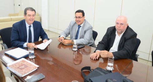 #PraTodosVerem: Ao redor de uma grande mesa de reuniões de vidro, 3 homens participam de uma reu...