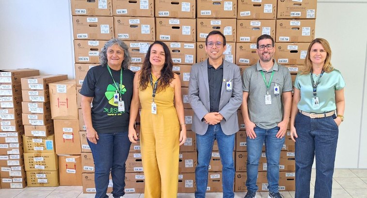 #PraTodosVerem: Na fotografia aparece um grupo de pessoas em frente a várias caixas empilhadas c...