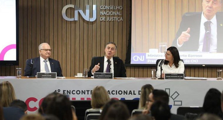 TRE-PB encontro nacional comunicação CNJ 1 #ParaTodosVerem: Três pessoas sentadas à mesa de trabalho em um auditório com várias outras pess...