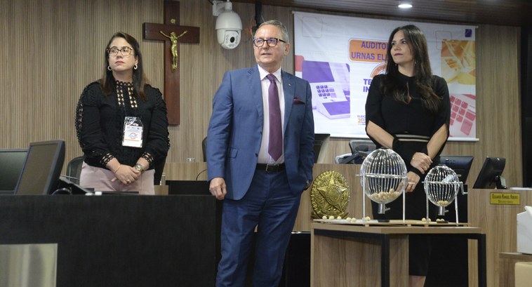 TRE-PB escolhe urnas eletrônicas que serão auditadas no 2º Turno das Eleições 2022 #ParatodosVerem: Três pessoas em pé, realizando um sorteio, são duas mulheres e um homem no centro.