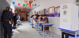 #PraTodosVerem: Na fotografia aparece um corredor com pessoas sentadas numa mesa e uma cabine el...