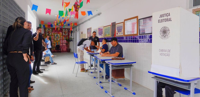 TRE-PB Estudantes de Remígio são eleitos para Câmara Mirim de vereadores #PraTodosVerem: Na fotografia aparece um corredor com pessoas sentadas numa mesa e uma cabine el...