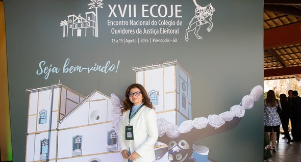 Foto 1: #PraTodosVerem: Na fotografia aparece uma mulher, usando óculos, em pé em frente a um pa...