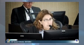 TRE-PB massaranduba #ParaTodosVerem: Painel retangular fotografia em plano médio de uma mulher sentada por trás de u...
