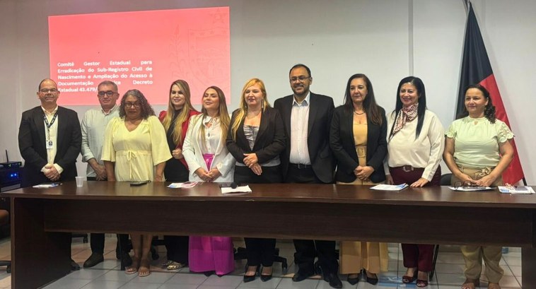 TRE-PB participa de evento de inclusão cidadã por meio da documentação básica #PraTodosVerem: Na fotografia aparece um grupo de pessoas em pé por trpas de uma mesa