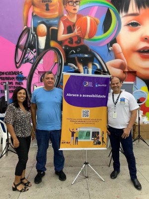 #PraTodosVerem: Três pessoas posam sorrindo ao lado de banner sobre acessibilidade, em frente a ...