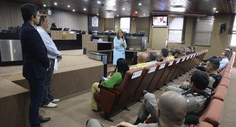 TRE-PB realiza segunda reunião voltada à segurança das Eleições 2024 #ParaTodosVerem: Três pessoas de pé e outras muitas sentadas ouvindo uma mulher falar ao microfone.