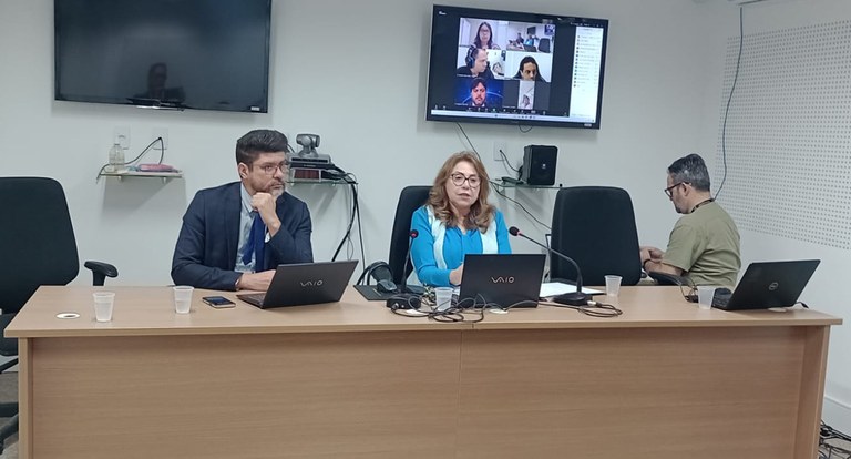 Painel retangular com três pessoas sentadas à mesa de trabalho, sendo uma mulher e dois homens, ...