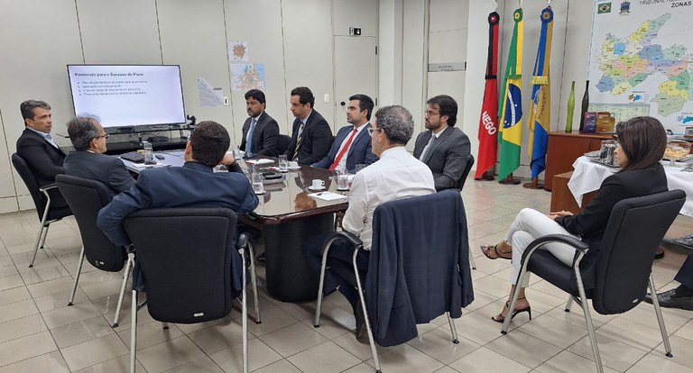 TRE-PB - Pessoas reunidas em volta de uma mesa, vendo um expositor eletrônico ao fundo.