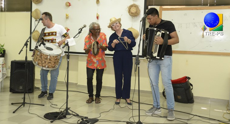 TRE-PB sextapontocom Escurinho #ParaTodosVerem: Painel retangular com quatro pessoas de pé, tocando instrumentos musicais, send...