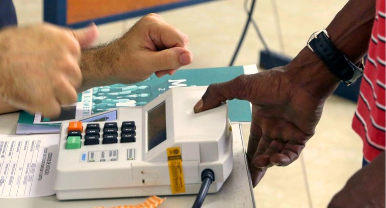 #PraTodosVerem: Na fotografia uma mão fazendo a identificação biométrica na mesa de votação da s...