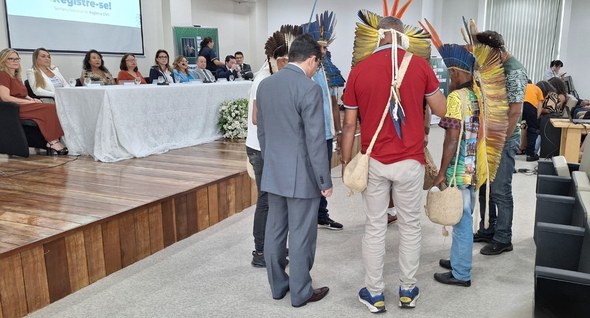 #PraTodosVerem: Na fotografia aparecem indígenas se apresentando a frente de uma mesa de honra, ...