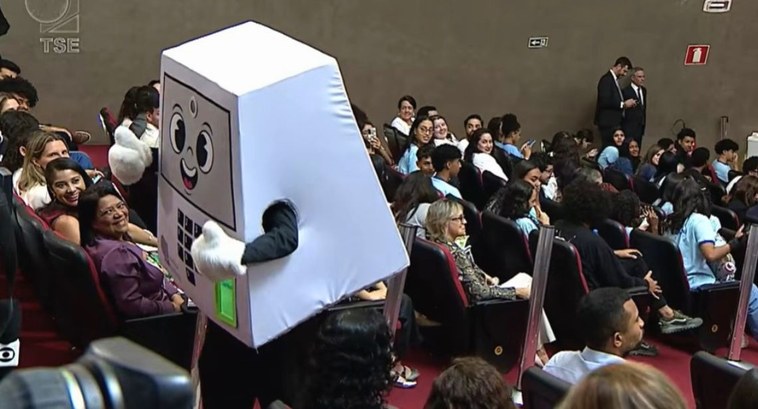 #PraTodosVerem: Na fotografia, aparece um mascote da urna eletrônica caminhando num auditório te...