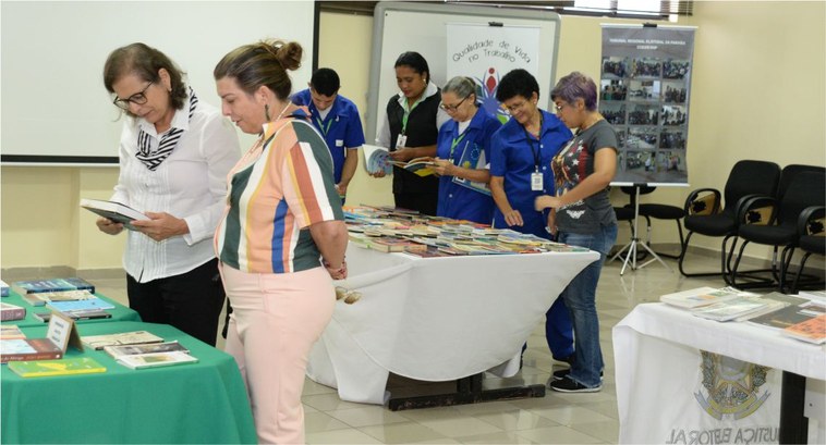 #PraCegoVer: Na fotografia aparecem vários participantes da Feira de Troca de Livros em volta da...